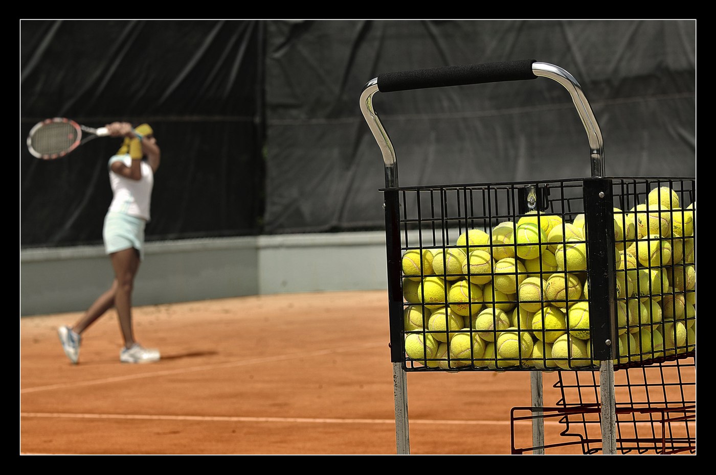 Joueuse de tennis sur terre battue, avec un panier de balles au premier plan.