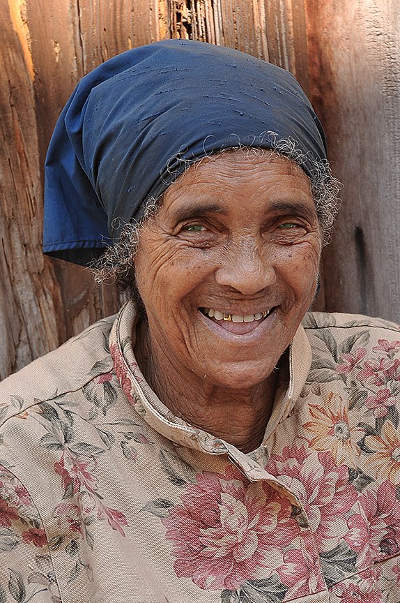 Femme souriante, coiffée d’un foulard bleu, devant une porte en bois.